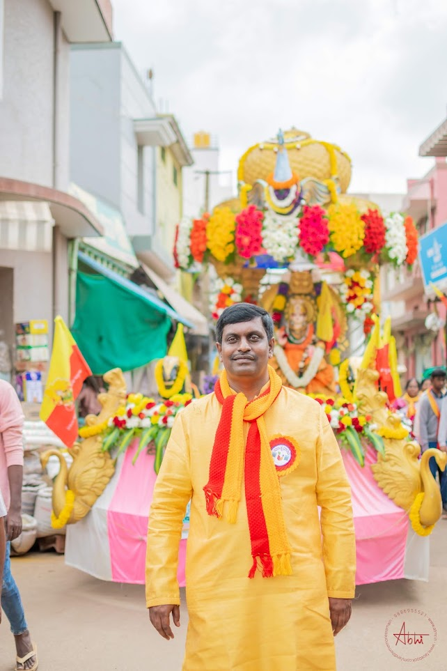 A volunteer in a yellow kurta before a decorated festival float during the Rajyotsava celebrations